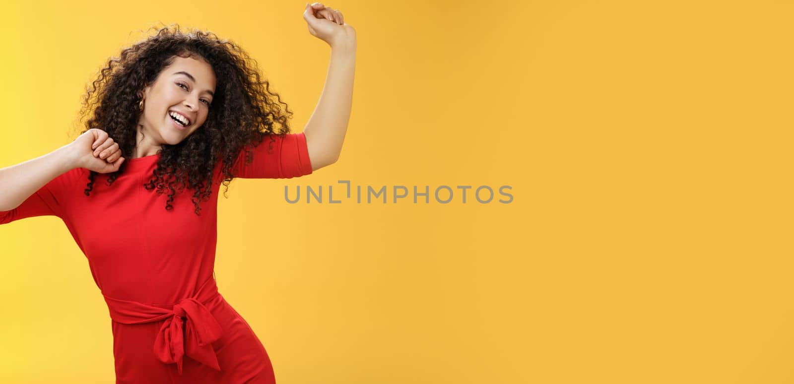 Carefree girl feeling uplifted and joyful dancing in red dress raising hands up happily tilting head and smiling broadly at camera as enjoying vacation, celebrating holidays over yellow background by Benzoix