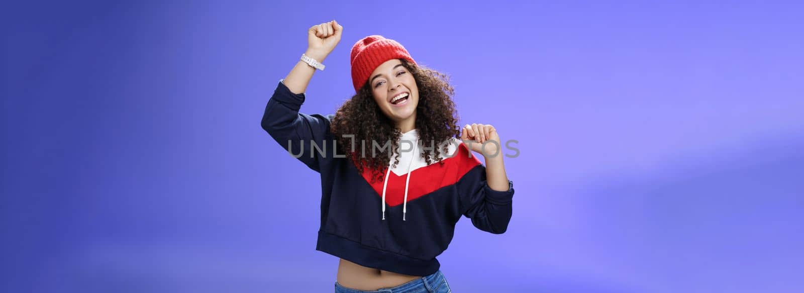 Hey come on dance with me. Friendly-looking bright and stylish cheerful woman with curly hairstyle wearing warm beanie raising hands up as enjoying great day, having fun outdoors over blue wall.