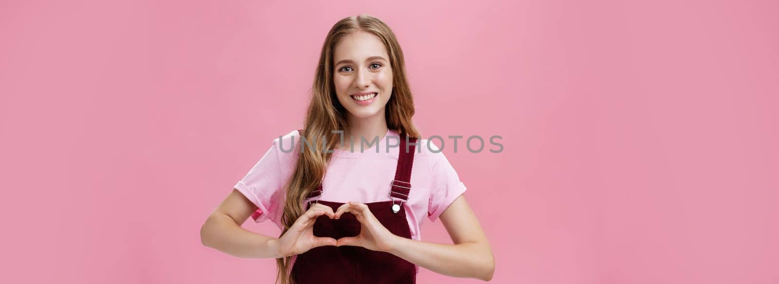 Girl loves family. Kind charming young woman in overalls with small tattoo on arm showing heart gesture over body and smiling lovely at camera expressing tender and cute attitude over pink wall.