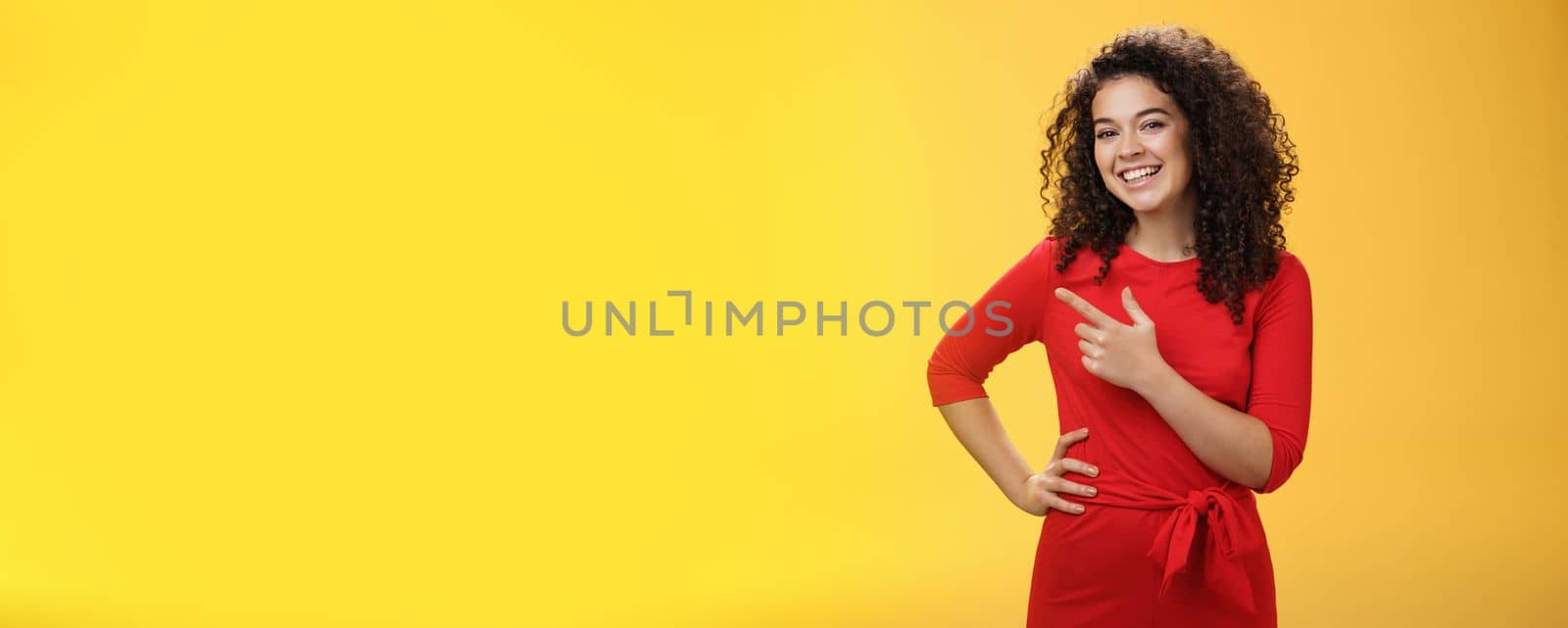 Portrait of friendly and assertive charming female coworker showing around newbie pointing left and smiling, laughing at camera having fun posing with hand on hip over yellow background by Benzoix