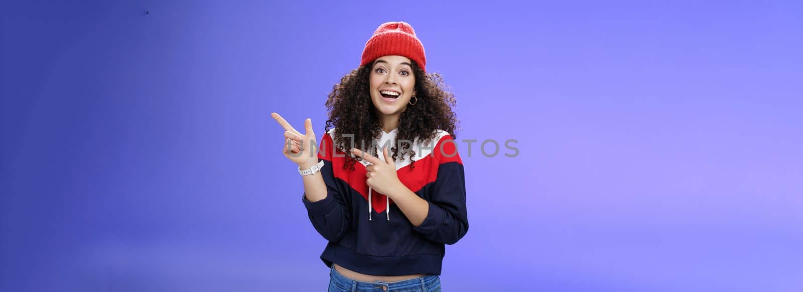 Lifestyle. Enthusiastic friendly-looking attractive young 20s woman with curly hair in warm beanie and sweatshirt smiling open mouth amazed and delighted as pointing at upper right corner over blue background.