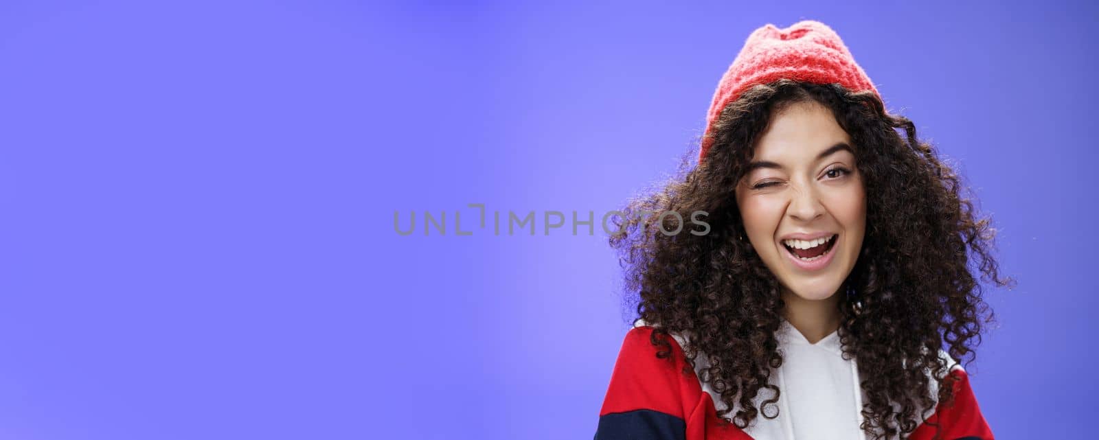 Charming cute woman hinting to go play snowballs with her winking joyfully as inviting us, smiling broadly wearing stylish red beanie and sweatshirt as posing against blue background.