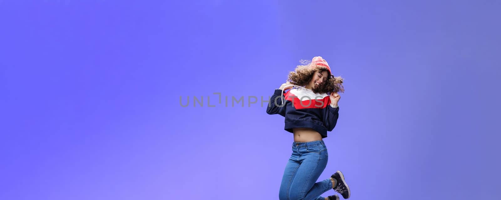 Girl jumping from happiness and delight feeling carefree close eyes and smiling broadly enjoying moment having fun raising hands being dreamy and playful over blue background. Lifestyle, fashion and positive emotions concept