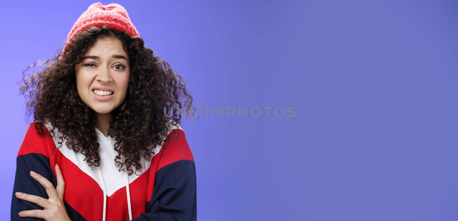 Woman with curly hair in winter beanie feeling uncomfortable and discomfort clenching teeth and frowning intense as hugging herself insecure and awkward, unwilling to say cruel rejection, feel awkward by Benzoix