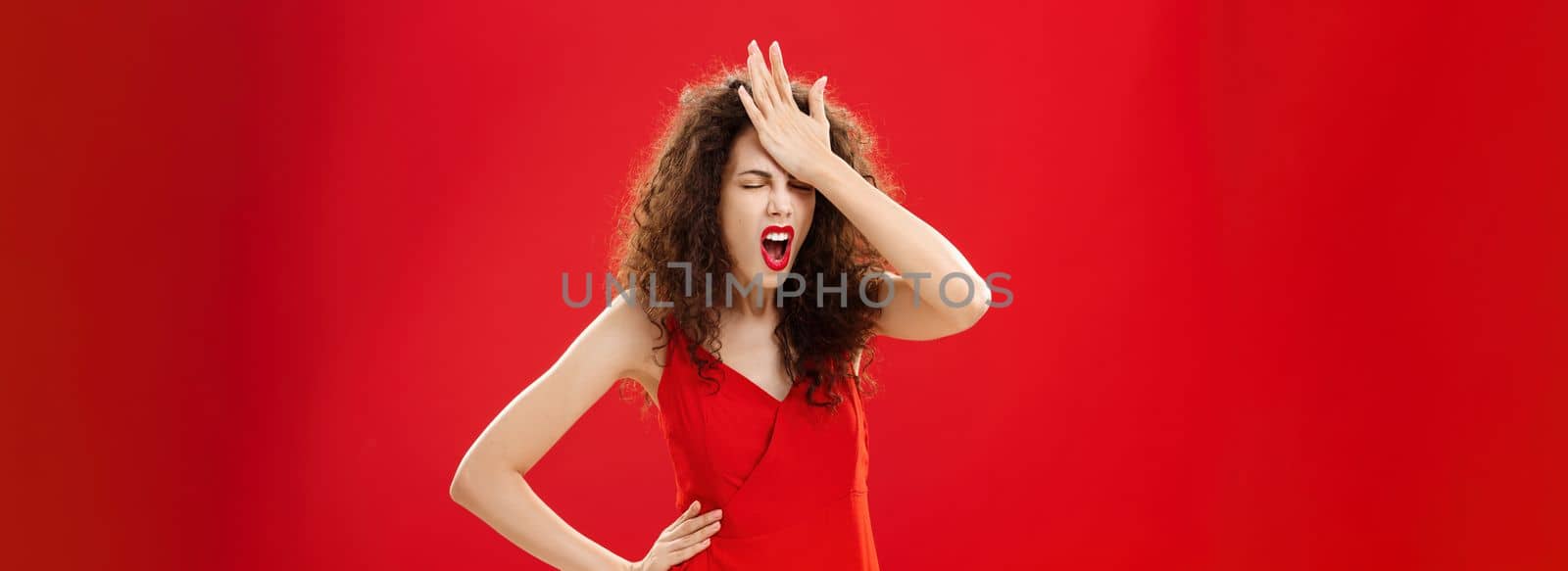 Woman being pissed with dumb people. Irritated forgetful adult caucasian female with curly hair in red elegant dress punching forehead with palm closing eyes and swearing recalling important thing by Benzoix