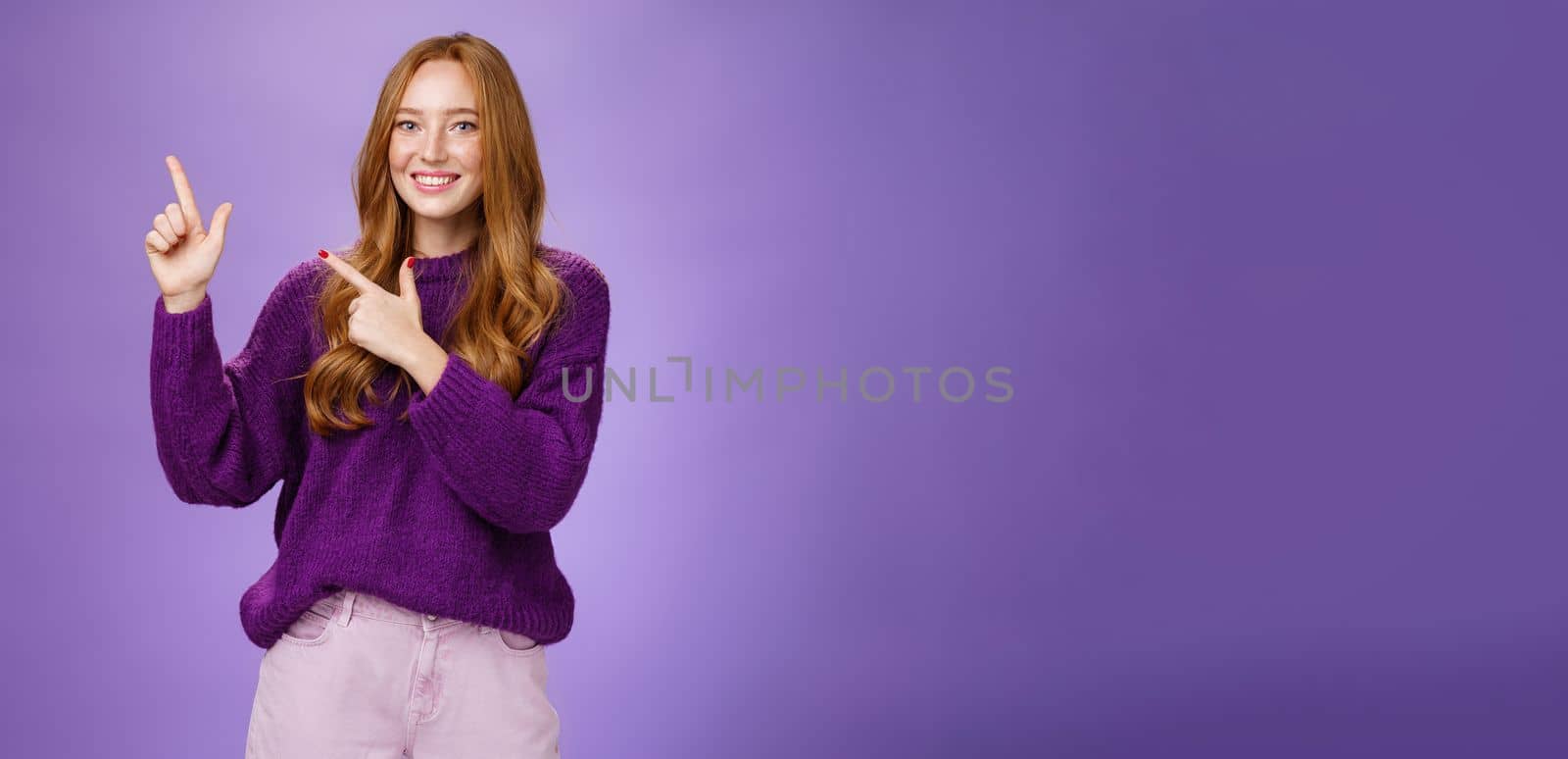 Charming assertive female shop assistant pointing at upper left corner to promote cool product smiling broadly feeling joyful and excited expressing friendly attitude as posing in purple sweater by Benzoix