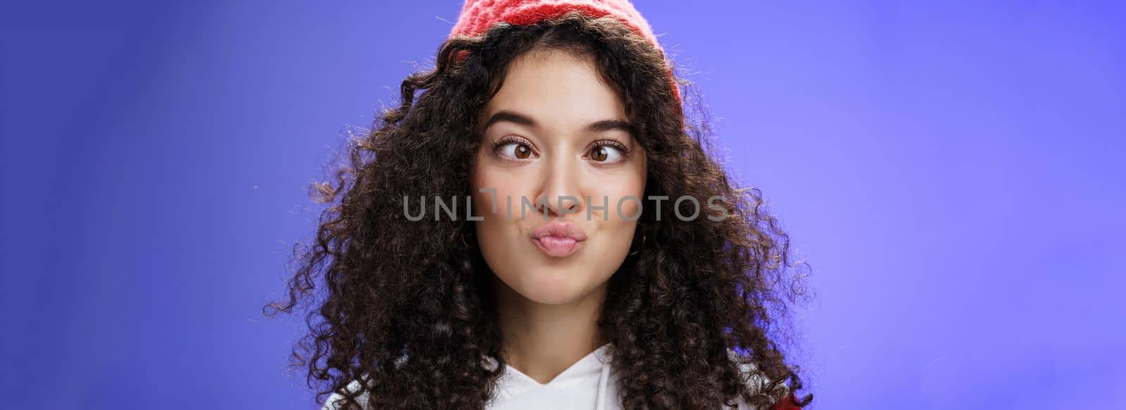 Headshot of funny and playful girl fooling around making faces not scared being funny folding lips like duck and squinting ice, aping standing childish and hilarious over blue wall in winter hat.