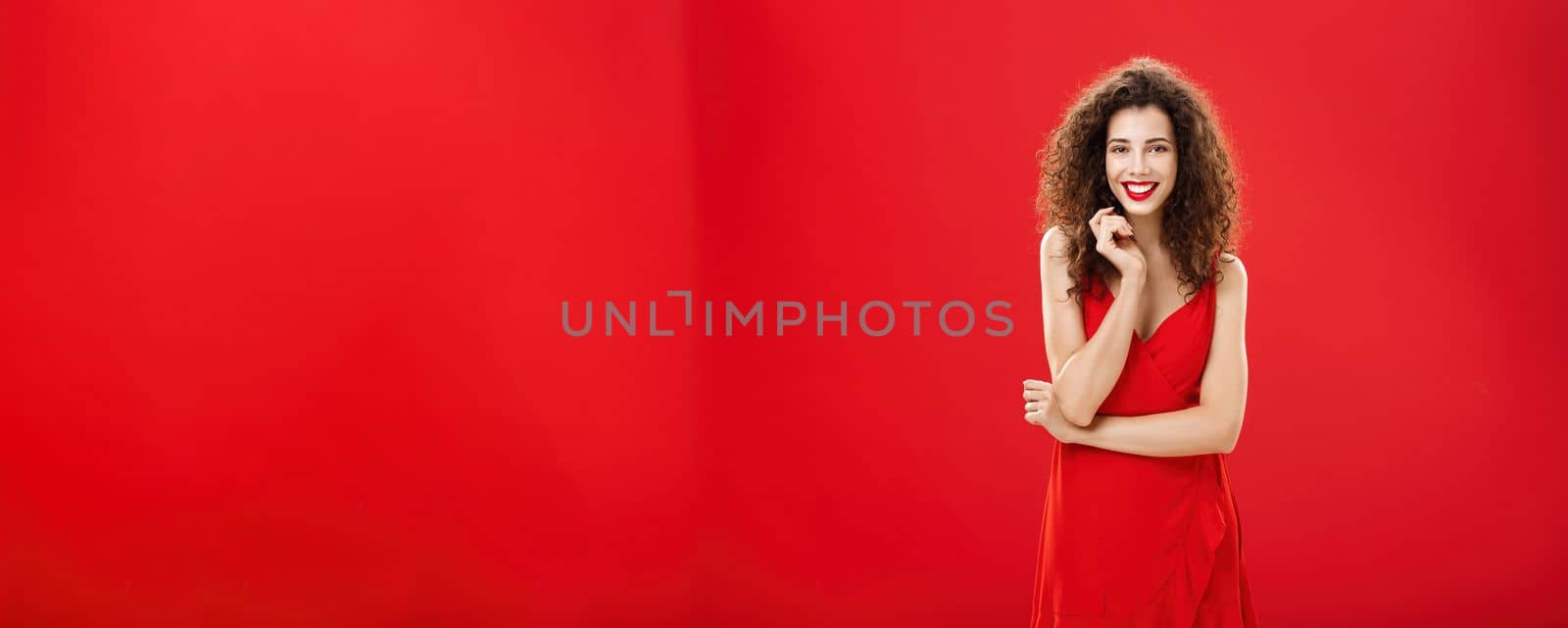 Passionate charming european woman over red background in elegant dress with curly hairstyle smiling cute, feminine playing with hair strand standing timid and silly, talking to person she admires by Benzoix