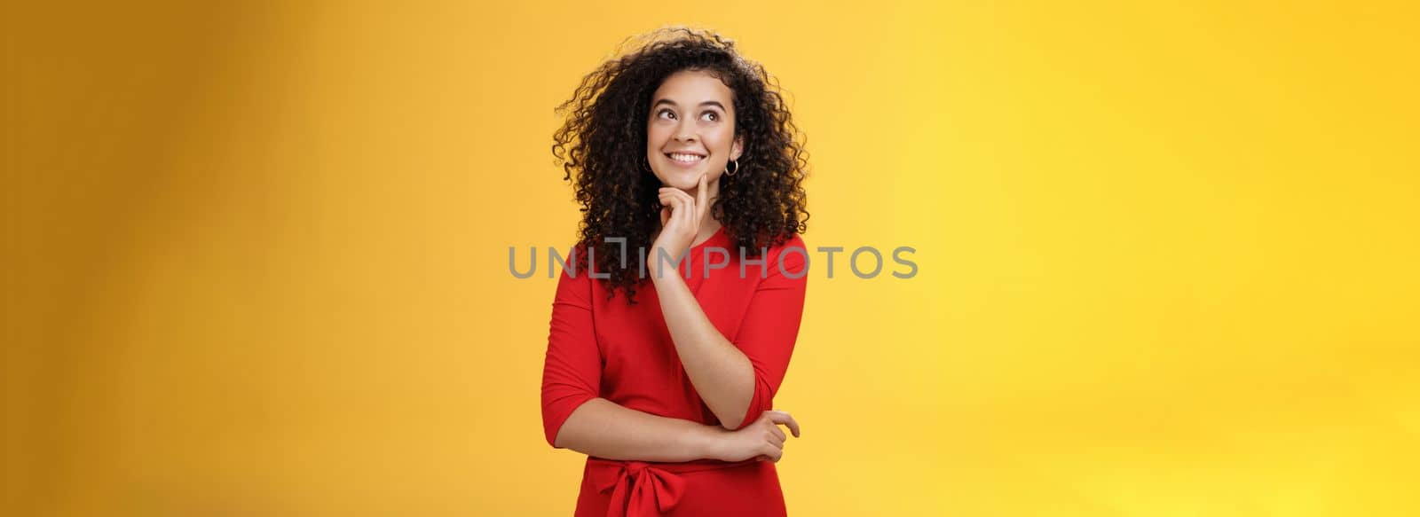 Girl having interesting idea, smiling as feeling confident plan might work. Charming happy woman with curly hair in red dress gazing at upper left corner thoughtful, thinking over yellow wall.