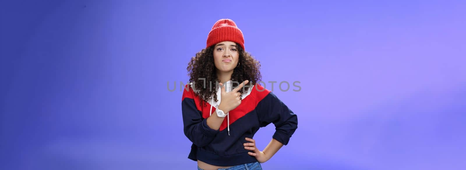 Girl feleing sad missing good chance, such shame. Portrait of displeased woman regretting frowning and pursing lips gloomy pointing at upper left corner upset wearing warm beanie over blue wall by Benzoix