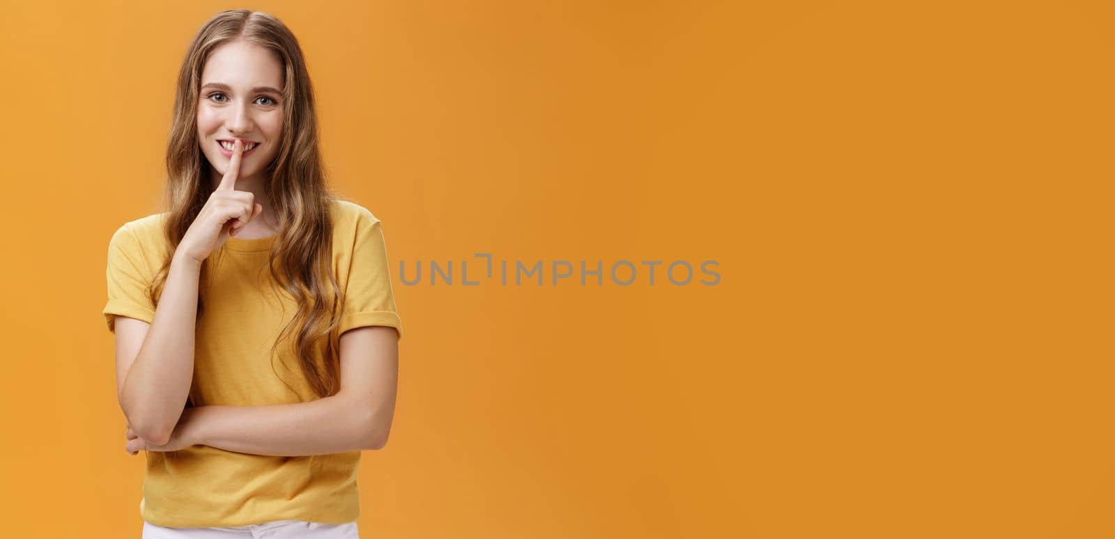 Girl wants keep secret betweet us. Portrait of charming good-looking slim pretty and young woman with long wavy hair and tattoo showing shh gesture with index finger over mouth against orange wall.