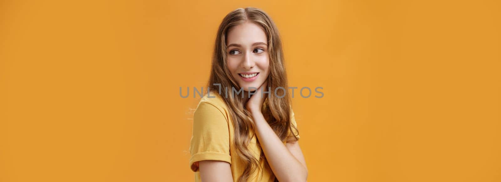 Soft and tender beautiful young girl with wavy fair hair turning back and look right touching neck gently smiling silly and sincere feeling pretty and self-assured posing over orange background by Benzoix