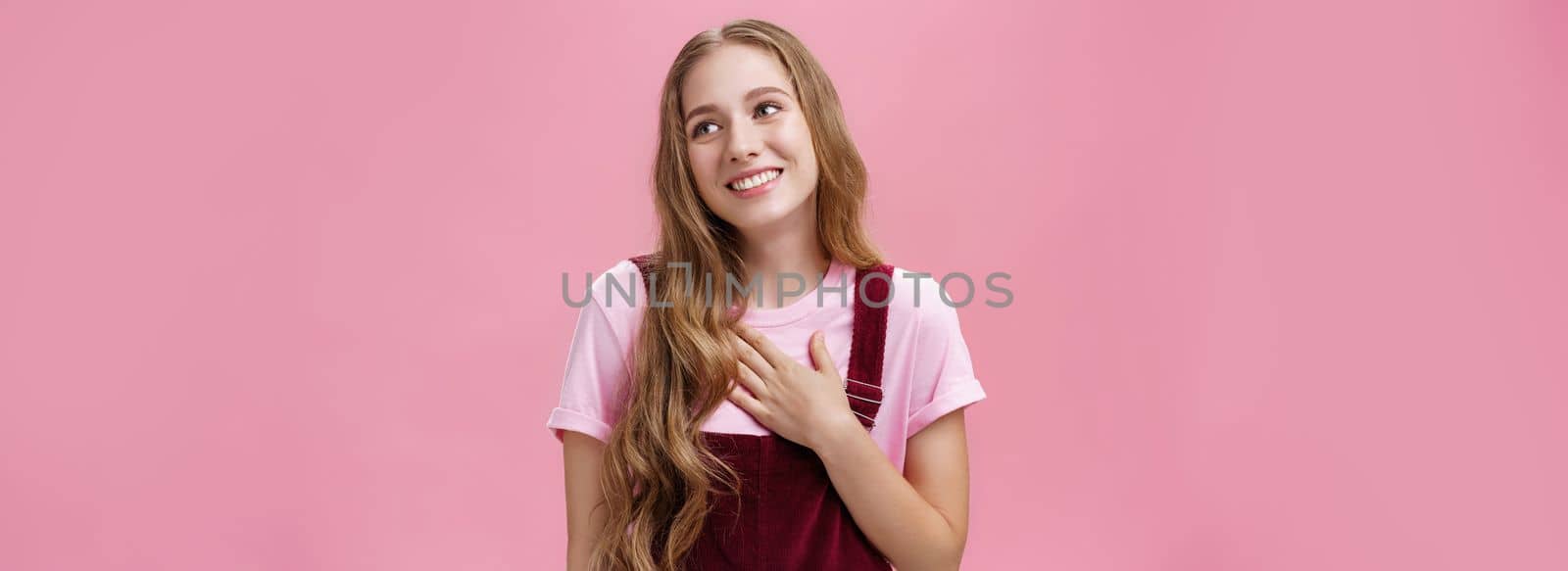 Waist-up shot of delighted charming friendly-looking tender female with cute white smile in corduroy overalls holding palm on breast and gazing right thankful, being grateful for compliments. Body language concept