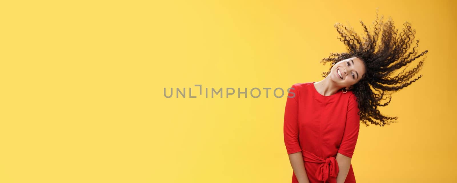 Bright happy and carefree playful woman waving curly hair making wave and smiling broadly as standing joyful in red dress over yellow background in good mood for future adventures by Benzoix