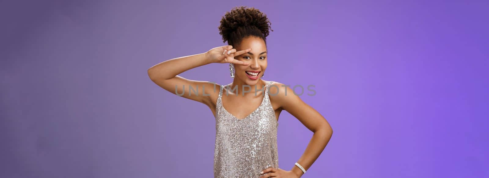 Joyful charming african-american woman in silver shiny evening dress smiling lively entertained grinning show victory peace gesture hold hand waist enjoying awesome party, blue background.