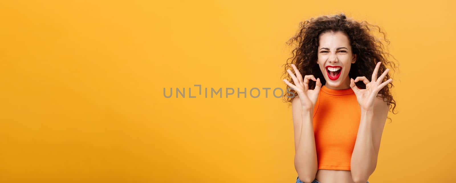 Charismatic happy and joyful attractive young female with curly hairstyle showing okay gestures with both hands in approval or confirmation smiling and laughing happily over orange wall by Benzoix