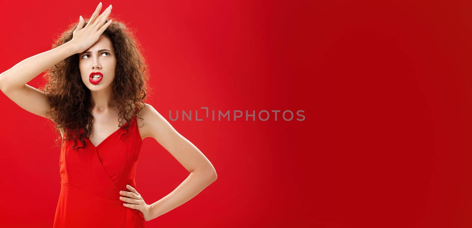Woman feeling irritated of stupid waiter messing up order punching forehead from annoyance rolling eyes up in anger frowning, swearing saying cursing words displeased standing in red dress. Emotions and body language concept