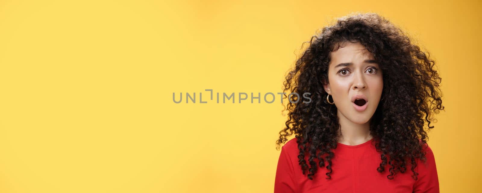 Lifestyle. Confused and frustrated young questioned woman with curly hair open mouth and raising eyebrow in surprise being displeased with unfair situation standing clueless and upset over yellow background.