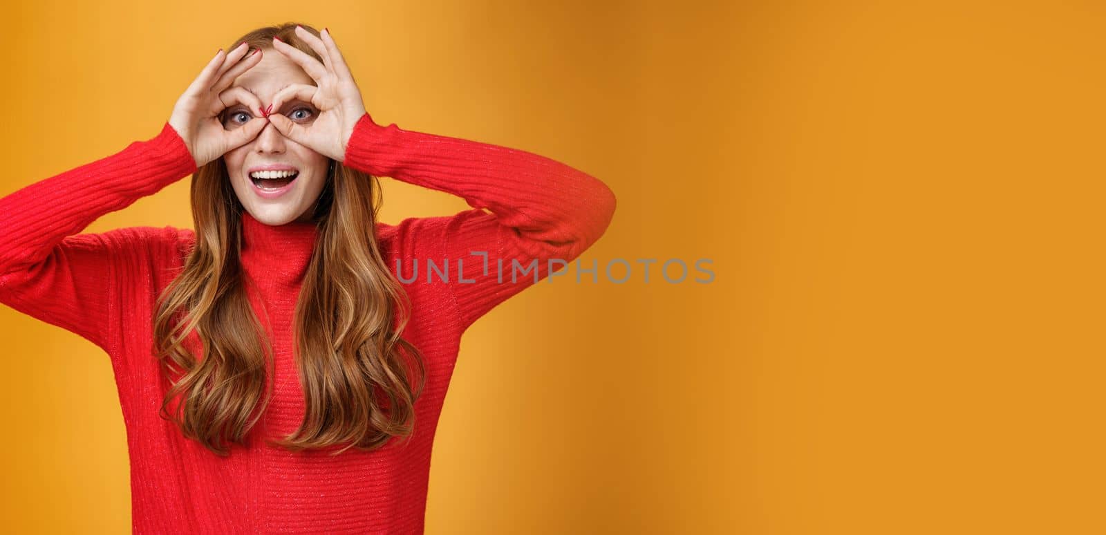 Waist-up shot of childish and playful funny ginger girl in red knitted dress making faces showing circles over eyes like goggles and smiling broadly having fun against orange backgound like child. Copy space