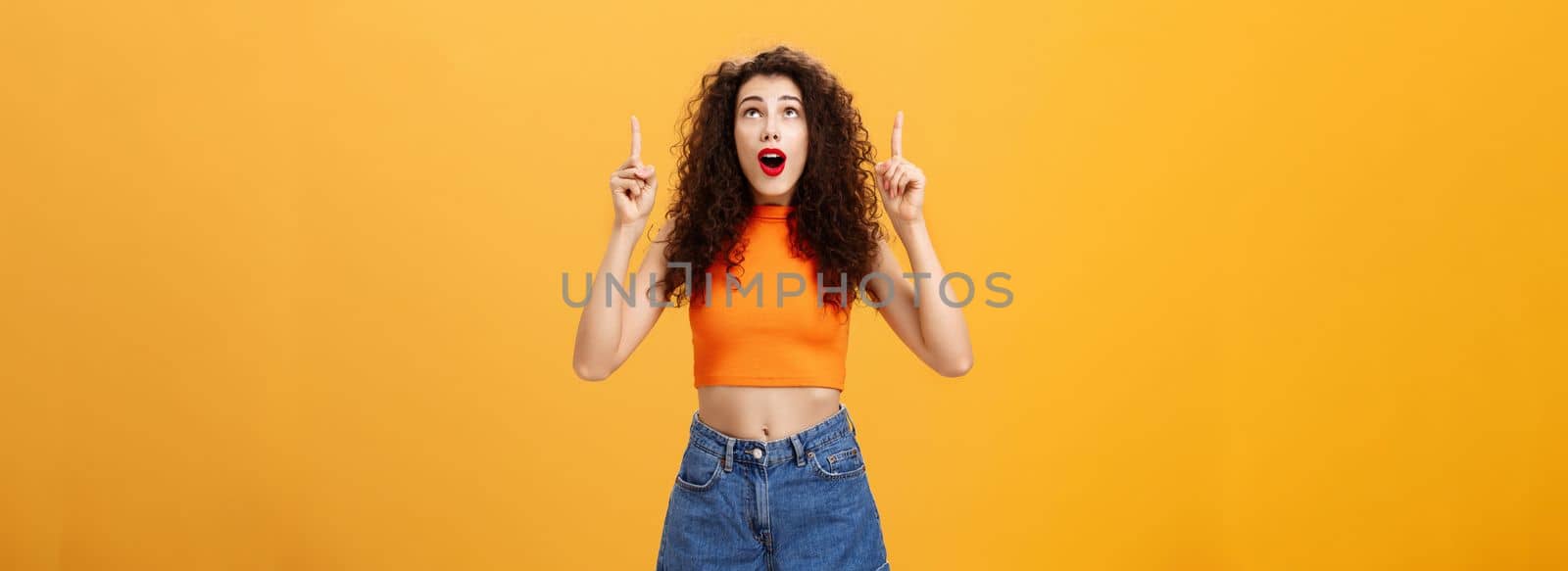Portrait of amazed emotive and charismatic young curly-haired woman with red lipstic looking and pointing up mesmerized and astonished standing under impression over orange background. Copy space