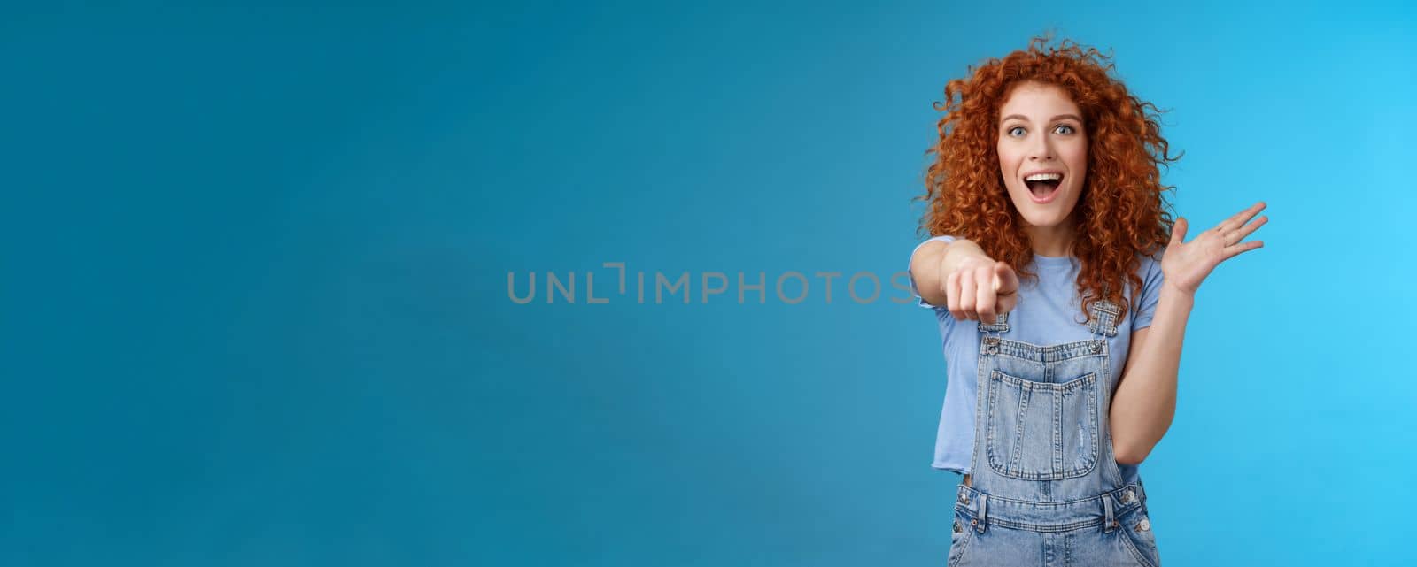 Excited impressed surprised cute redhead european woman curly astonished happily pointing index finger camera gesturing lively thrilled see awesome thing reacting amazed standing blue background by Benzoix