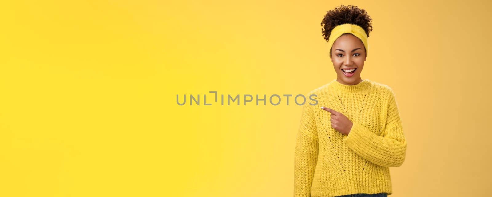 Friendly good-looking enthusiastic black charming girl in headband sweater smiling broadly amused pointing left awesome place showing you perfect blank space advertisement, posing yellow background.