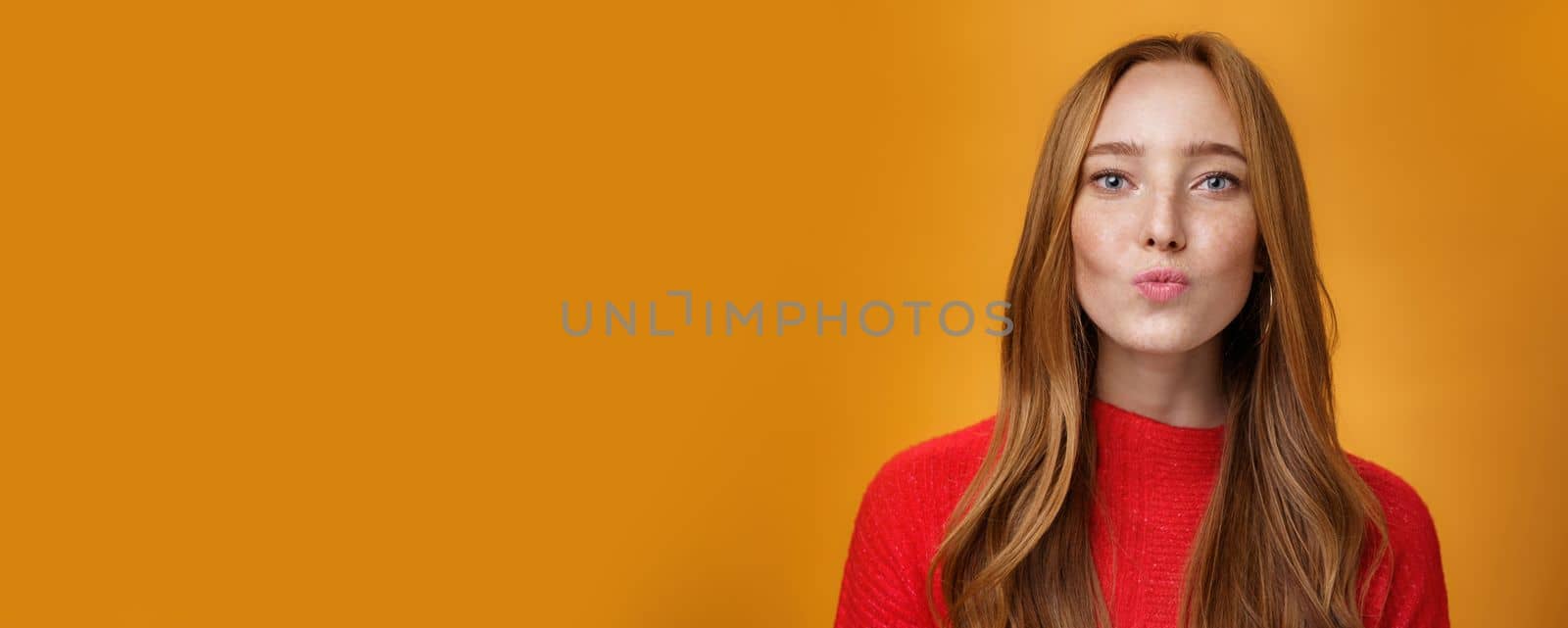 Close-up shot of flirty good-looking sensual redhead female in red knitted clothes folding lips in kiss and gazing at camera as giving mwah feleing romantic and pretty against orange background. Body language, fashion and people concept