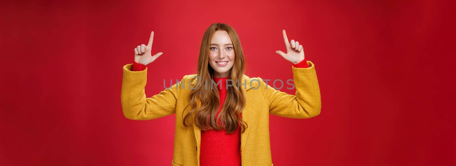 Indoor shot of redhead attractive woman in yellow fall coat raising hands promoting advertisement as pointing up and smiling broadly with satisfied pleasant expression over red background by Benzoix