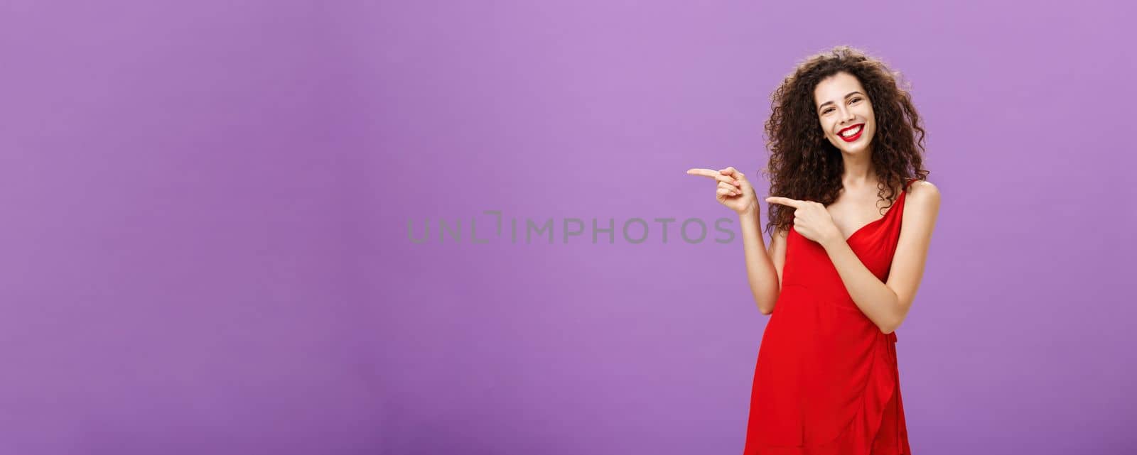 Girl suggesting copy space to try. Portrait of good-looking elegant caucasian woman with curly hair in stylish evening red dress pointing left and smiling inviting customers over purple background.