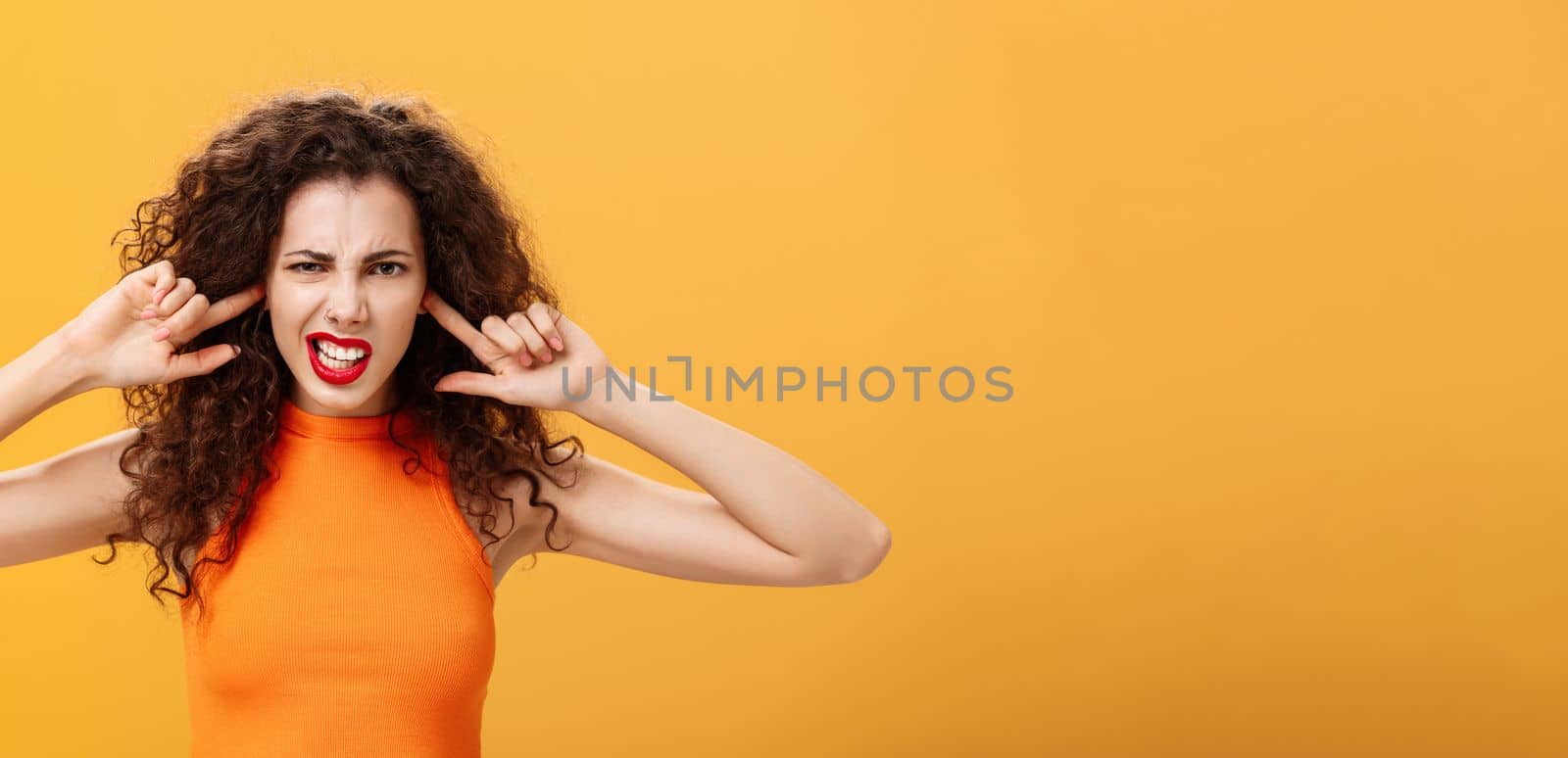 Waist-up shot of irritated bothered stylish woman with curly hair and red lipstick squinting clenching teeth waiting for loud bang covering ears with index fingers being annoyed of noisy neighbours by Benzoix