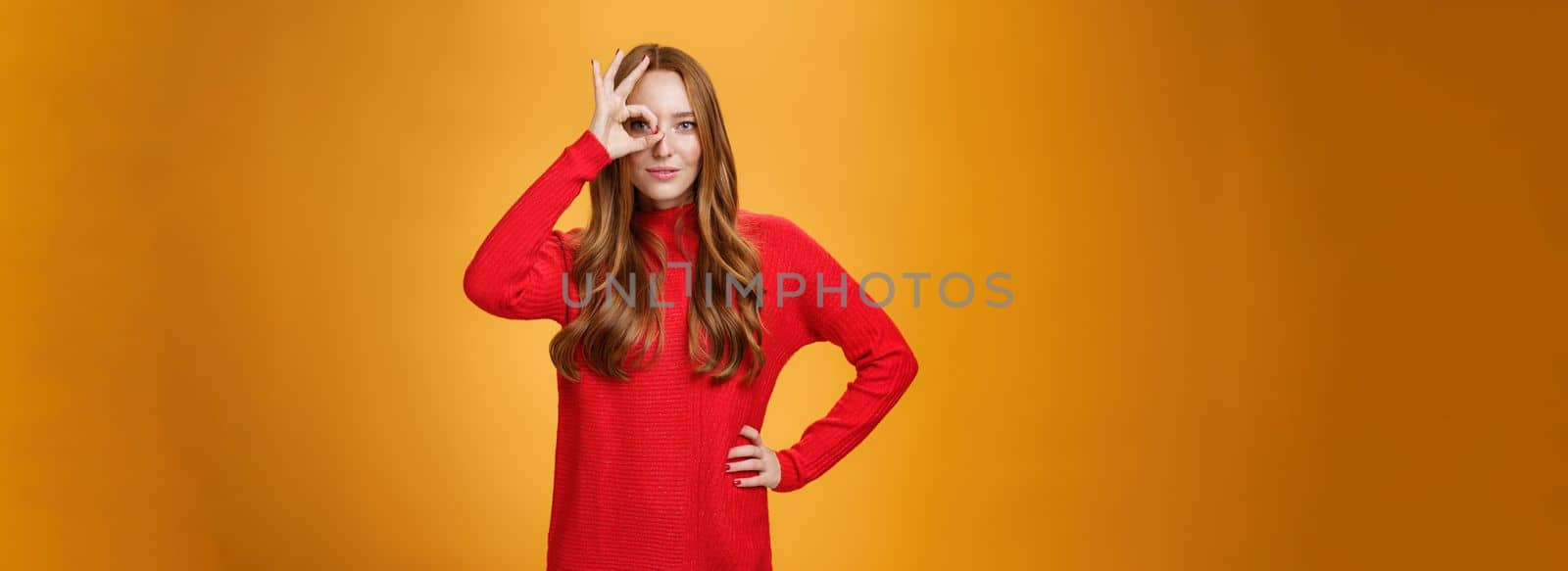 I got you on aim. Self-assured expressive and confident attractive redhead woman in red warm dress showing okay or zero sign on eye and peeking through as smiling satisfied over orange wall.