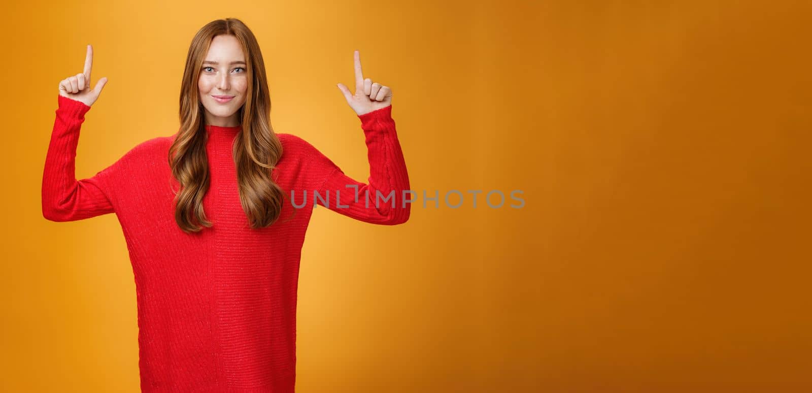 Look up you not regret it. Portrait of confident and stylish young redhead woman in 20s wearing knitted red dress pointing upwards and smiling assertive with self-assured grin over orange background by Benzoix