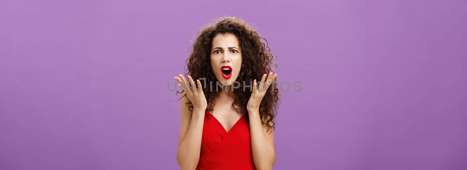 Rich arrogant and snobbish european woman. with curly hairstyle in red evening dress arguing with made frowning looking confused and displeased shaking palms in disappointment posing over purple wall.