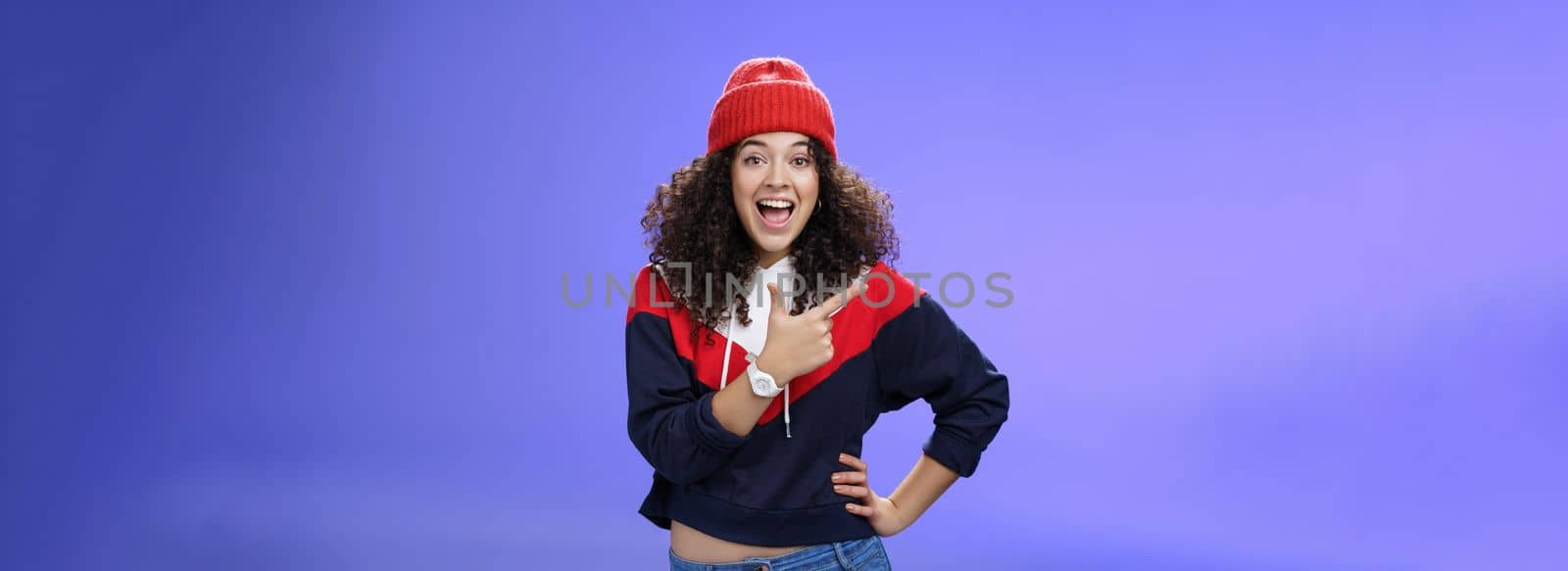 Portrait of enthusiastic and sociable european woman in warm hat and sweatshirt smiling delighted with amused grin as pointing at upper left corner impressed and astonished with awesome promotion by Benzoix