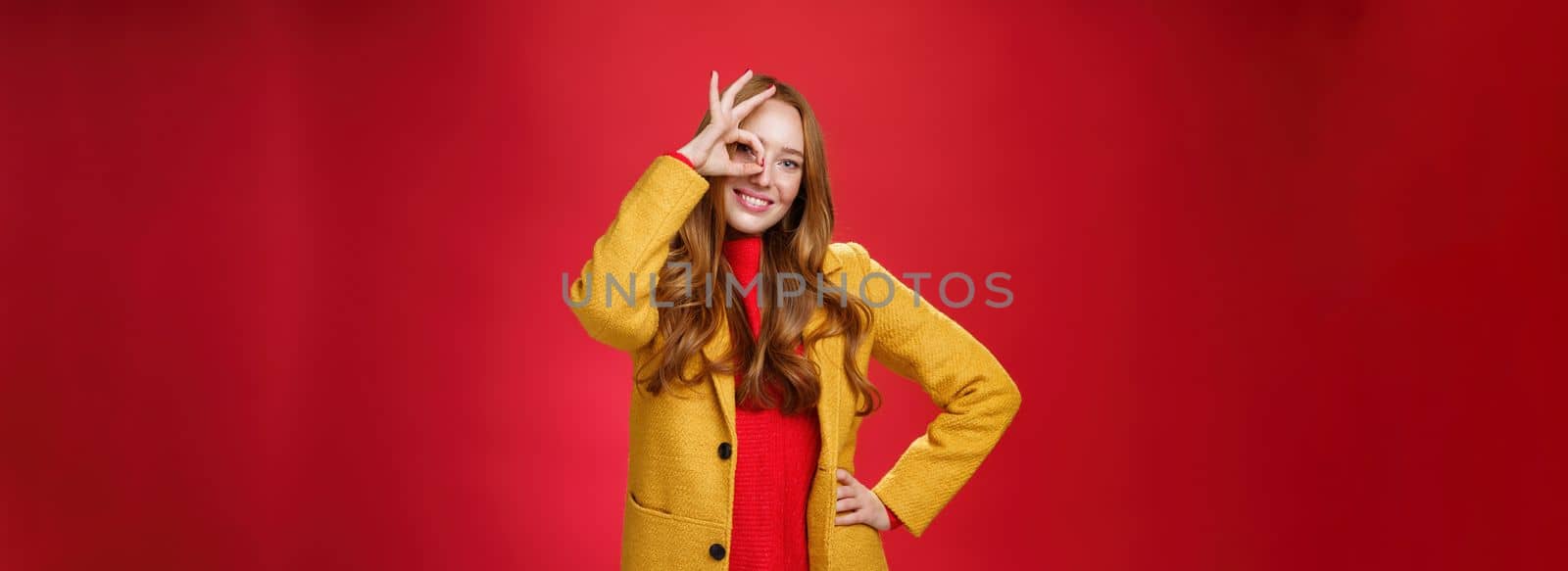 Bright and sunny girl feeling happy even on rainy days standing in yellow stylish coat over red background showing okay gesture over eye, smiling broadly at camera, holding hand on waist by Benzoix