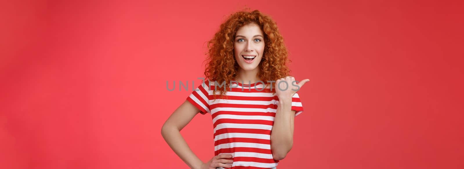 Lifestyle. Sassy good-looking emotive happy smiling redhead european female curly hairstyle pointing thumb left grin assertive cheeky hold hand waist directing promo advertising offer red background.