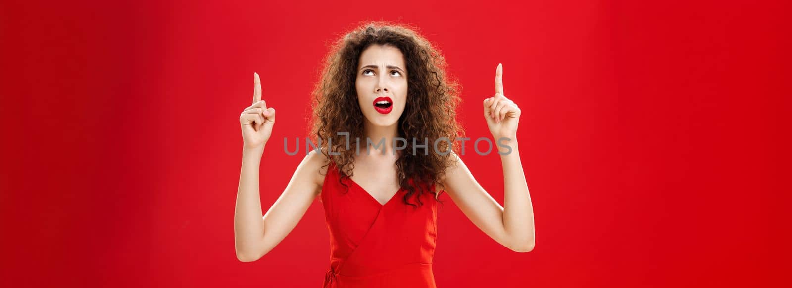 Troubled woman cannot understand what happening. Portrait of clueless silly european female with curly hair in red dress looking and pointing up perplexed and questioned posing over studio background. Copy space
