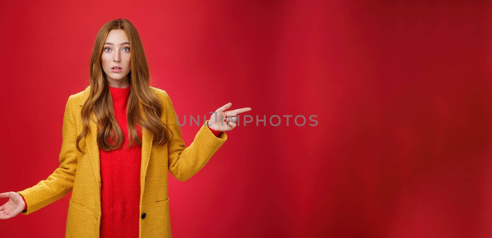Confused and questioned cute redhead girl in yellow autumn coat pointing right as shrugging making clueless gesture with hand open mouth wondered as asking question over red background by Benzoix
