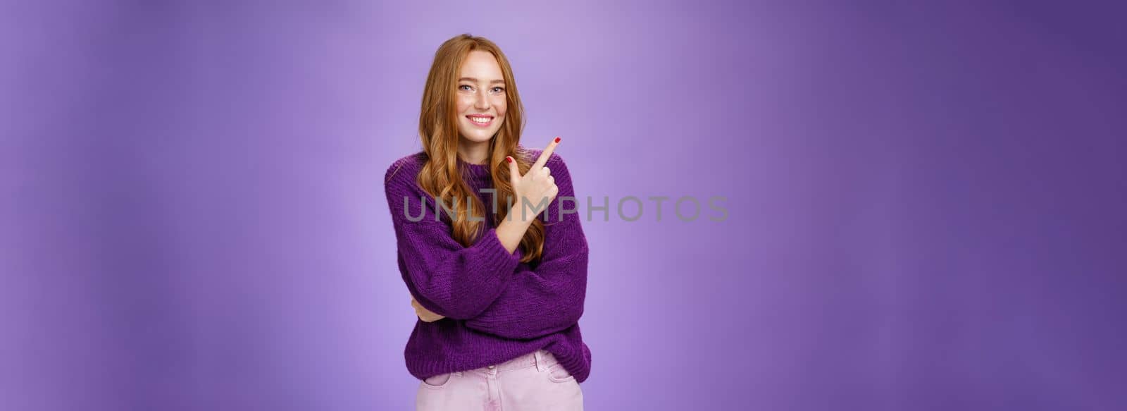 Lifestyle. Charming friendly-looking bright redhead woman with freckles and makeup in purple warm sweater pointing at upper left corner and smiling delighted and cute at camera as showing cool place hang out.