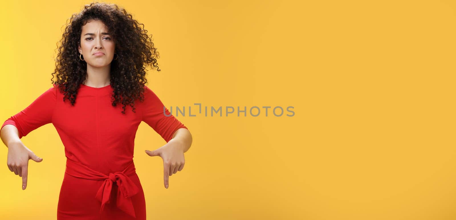Gloomy disappointed cute silly curly-haired female in red dress frowning upset and making sad smile as pointing down with regret and unsatisfied expression, jealous or displeased over yellow wall.