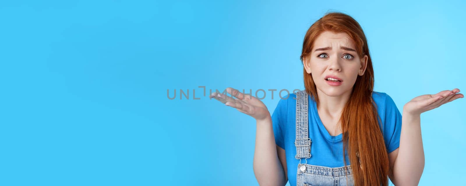 Perplexed uneasy stupified worried redhead woman shrugging, have no idea, frowning nervously, cannot understand what happened, stand clueless unsure, pose blue background by Benzoix