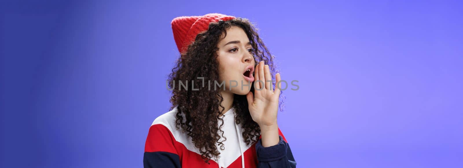 Portrait of worried sister calling sibling outdoors open mouth looking left seriously and holding palm near lips as shouting name searching someone, posing concerned over blue background.