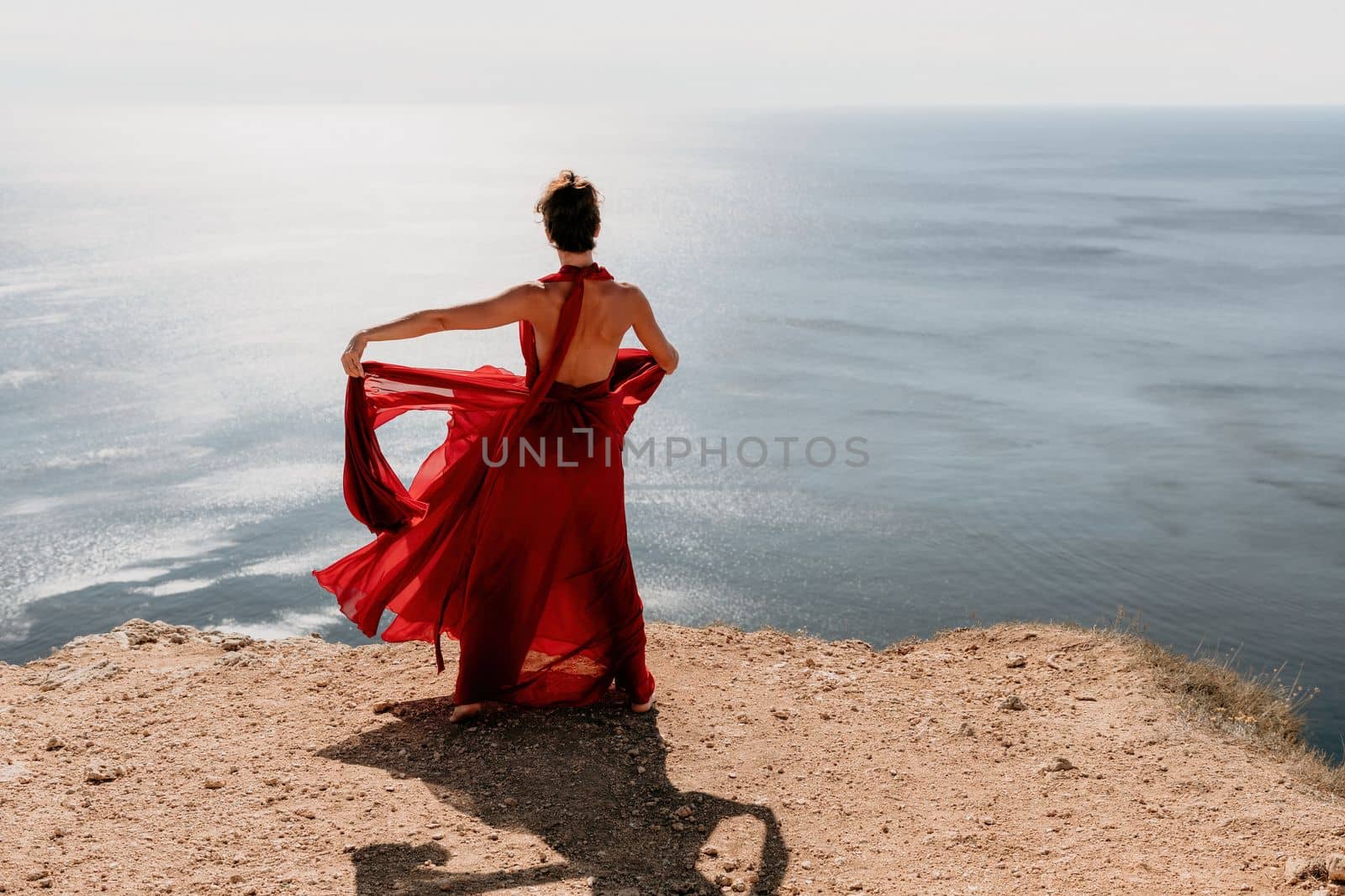 Side view a Young beautiful sensual woman in a red long dress posing on a rock high above the sea during sunrise. Girl on the nature on blue sky background. Fashion photo.