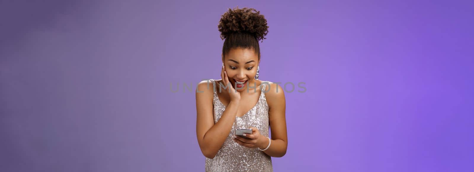 Surprised astonished young african american girl holding smartphone look display imressed super happy reading message touch cheek widen eyes gladly receive great news, standing blue background.