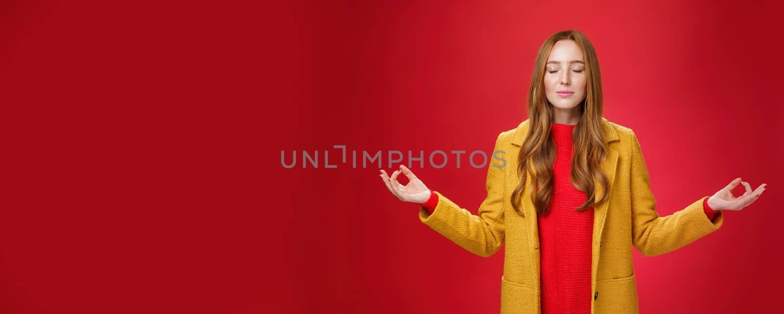 Girl keep calm releasing stress with meditation, posing in yellow coat, close eyes and looking relieved as extending hands sideways with mudra gesture, doing yoga against red background in lotus pose.