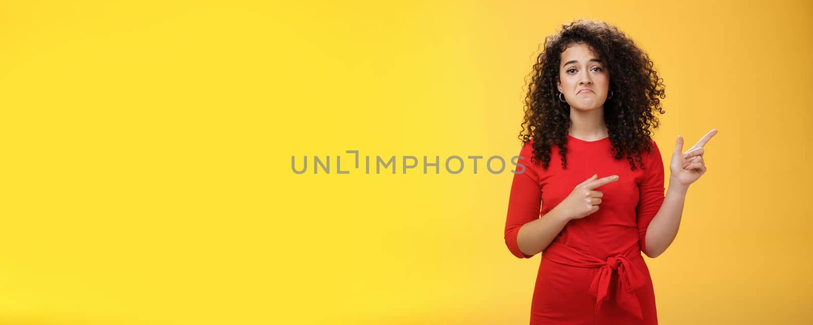 Waist-up shot of upset displeased girl with curly hair being down in dumps pointing at upper right corner with disappointed gloomy smile, feeling regret, sadness standing like loser over yellow wall.