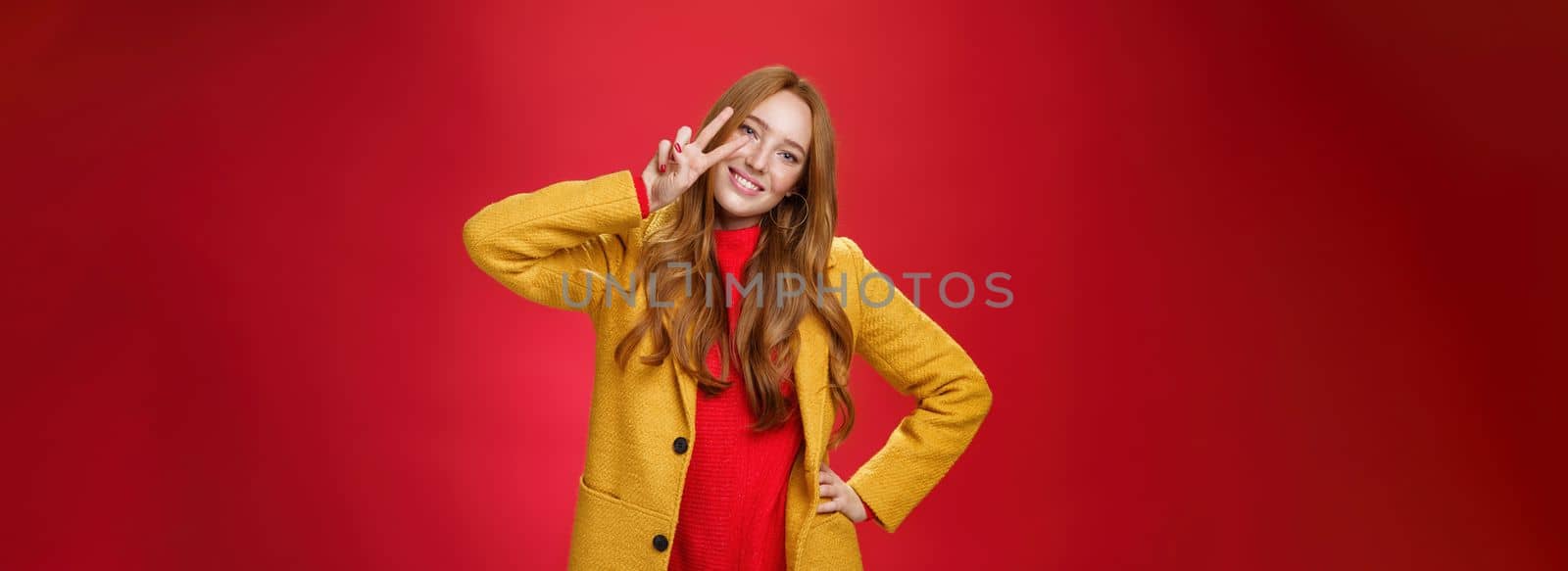 Friendly girl smiling happily, tilting head and showing peace or victory gesture near face, holding hand on hip as posing in stylish elegant yellow coat and sweater against red background. Style, emotions and fashion concept