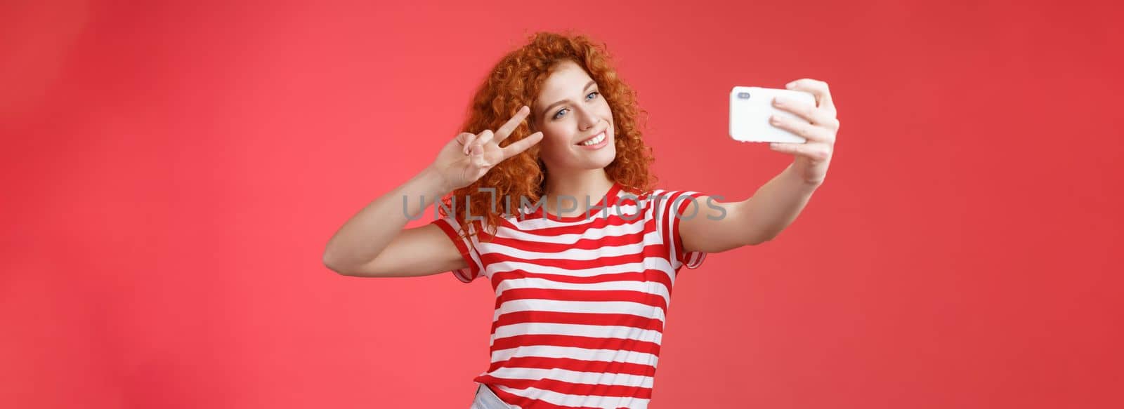 Stylish carefree good-looking redhead curly woman enjoy summer holidays show victory peace gesture eye tilt head cute record video hold smartphone taking selfie red background. Copy space