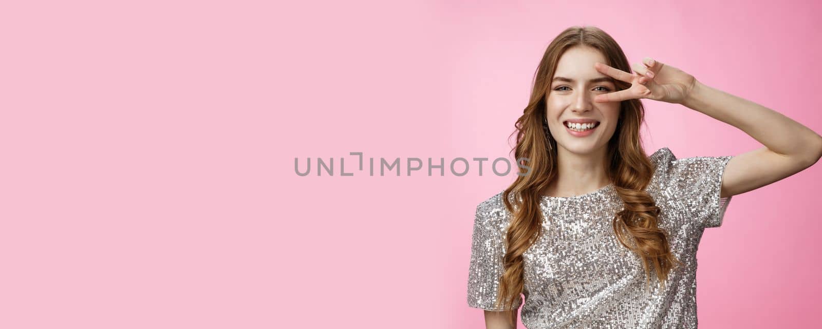 Positive carefree lucky charming glamour woman curly hairstyle party outfit having fun showing victory peace gesture dancing disco smiling broadly enjoying awesome atmosphere, pink background.