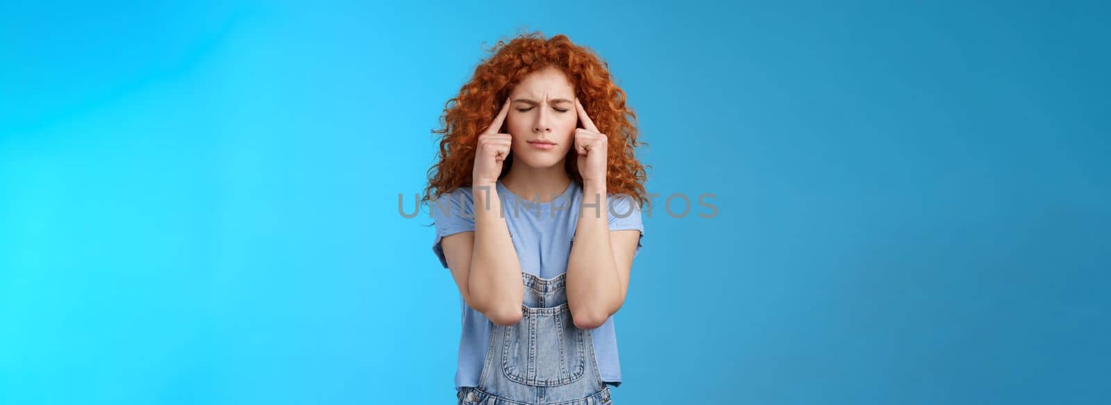 Come on girl think. Perplexed distressed concerned intense redhead curly woman close eyes frowning touch temples suffer migraine painful headache cannot concentrate blue background. Copy space
